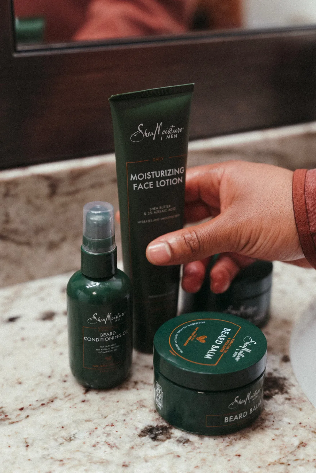 SheaMoisture Men beard oil, beard balm and face lotion on a bathroom counter, a hand reaching in